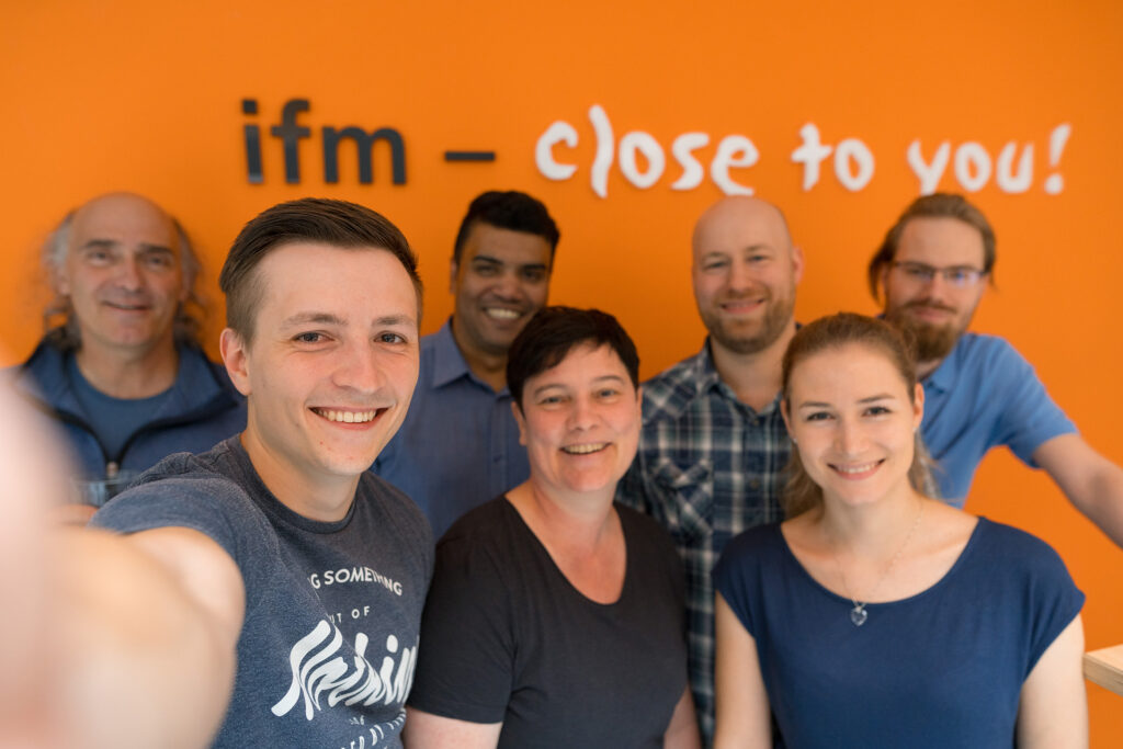 Mitarbeitende der ifm software machen ein Selfie vor dem ifm Logo